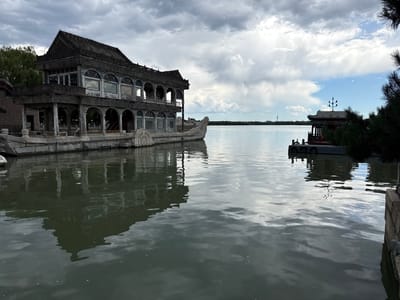 The image depicts the Marble Boat (Qingyanfang) on Kunming Lake within the Summer Palace in Haidian District, Beijing, China. A two-story stone structure designed to resemble a boat occupies the left and central midground. This structure features traditional Chinese architectural elements, including a tiled roof, ornate carvings, arched windows on the lower level, and rectangular windows on the upper level. Its right end terminates in a decorative scroll-like prow.
The water of Kunming Lake fills the foreground and midground, exhibiting a smooth, reflective surface with subtle ripples, mirroring the sky and architectural elements. To the far right, a smaller, traditional Chinese pavilion-like structure with a curved, multi-colored roof and a decorative lamppost on its peak is situated on a dock with circular black fenders. This pavilion is partially framed by dark green conifer foliage in the extreme right foreground.
The sky above is predominantly overcast with white and grey clouds, though distinct patches of blue sky are visible, particularly towards the distant horizon. A low treeline is present along the distant shoreline, and a slender communication tower (resembling the Central Radio and TV Tower) is discernible against the sky in the far distance on the right side. Green deciduous foliage, likely trees, is visible behind the Marble Boat on the left. Stone embankments line the immediate right edge of the water. No individuals are present in the scene.