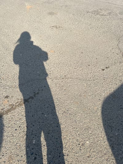 The image shows a patch of asphalt pavement. On the pavement is the shadow of a person and what appears to be the shadow of a hand holding a phone. The location is possibly Yugo-Kamskiy, Russia. The pavement has a rough texture. There is no obvious interaction in the scene. The environment is urban.