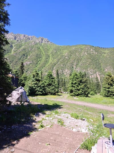 The image depicts a mountainous landscape with a clear blue sky. In the foreground, there is a large rock and a small, dark-colored structure partially visible. Several coniferous trees are scattered across the scene, with a dirt path winding through the grass and trees. In the background, a lush green mountain rises, its slopes covered in vegetation and rocky outcrops. On the far right, a black office chair with a metal frame is positioned on a wooden deck. The setting appears to be a natural, outdoor environment, possibly a campsite or a remote area.