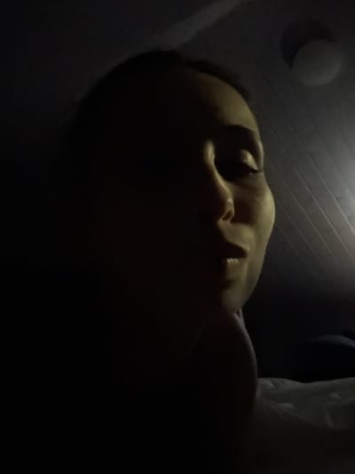 A low-angle, close-up, dimly lit indoor shot features a person's face from the side. The light source appears to be above and to the right, casting a strong shadow over the left side of the face. The person's right eye is visible, and their lips are slightly parted. The background consists of a ribbed ceiling and a circular light fixture. A white surface, possibly a bed or pillow, is visible in the lower right corner. The image is dark and somewhat blurry, making it difficult to discern fine details.