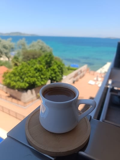 A close-up shot features a white coffee cup filled with dark coffee, placed on a light brown wooden coaster. The cup is positioned on the edge of a balcony or railing, with its handle visible.

In the background, a serene scene unfolds with a clear blue sky overhead. The turquoise waters of the sea stretch out towards a distant hazy landmass. Lush green trees and vegetation are visible, along with what appears to be a resort or residential area with buildings and a patio. Sunlight illuminates the scene, suggesting it is daytime with clear weather, possibly a warm and sunny day. The overall mood is one of relaxation and tranquility, perhaps a moment of quiet enjoyment with a coffee overlooking a beautiful coastal view in Urla, Turkey. There is no visible text in the image.