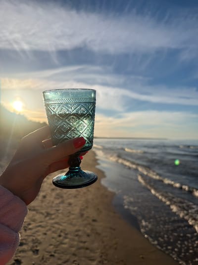 This is a horizontal, medium shot of a person's hand holding a decorative blue glass goblet at eye level. The hand, adorned with red nail polish, is gently cupping the stem of the goblet. The glass itself has an intricate diamond-patterned design etched into its surface, and it catches the sunlight, refracting it into a soft glow.

The background is a beach scene, with the sun setting or rising over the ocean. The sky is a pale blue, streaked with wispy white clouds. The ocean water is visible to the right, with small waves lapping at the sandy shore. The sand appears wet and dark where the water has receded, and lighter and drier further up the beach. The sun is low on the horizon, casting a warm, golden light across the scene and creating a lens flare.

The overall mood is peaceful and serene, suggesting a moment of quiet enjoyment or reflection by the sea. The time of day is likely late afternoon or early morning, given the low angle of the sun and the soft, warm light. The location is identified as Plieņciems, Latvia. No text is visible in the image.