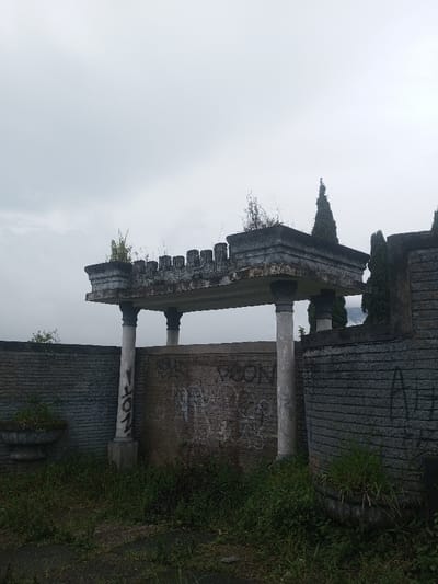 An overgrown, dilapidated stone structure under a gray sky. Columns support a decaying, castellated roof. Graffiti mars the central wall, framed by textured stone walls on either side. Overgrown vegetation covers the ground, obscuring the base of the structure and planters. Tall, narrow trees are visible in the background, adding to the somber, neglected atmosphere.