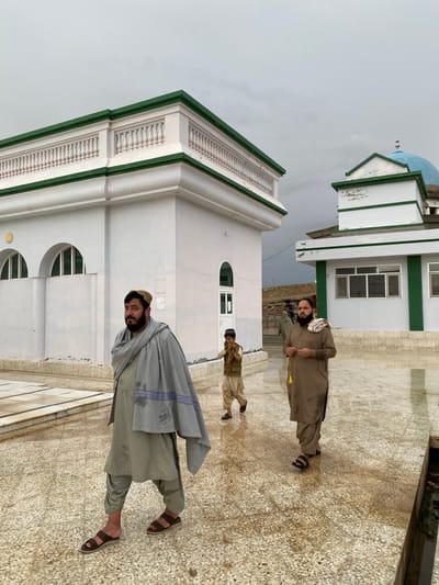 The image captures an outdoor scene at a building complex in Arghandab, Afghanistan, under an overcast sky. The ground is a wet, light-colored speckled pavement, indicating recent moisture. In the foreground, three individuals walk. On the left, a man with a dark beard and a light-colored cap wears a light olive-green shalwar kameez and a damp, grey shawl with a patterned dark blue border, walking in brown sandals and looking towards the viewer. A young boy, dressed in a light brown shalwar kameez and a dark cap, walks slightly behind the first man, holding a light-colored, indeterminate object in his left hand. On the right, a second bearded man in a damp, brown shalwar kameez holds a strand of yellow-tasseled prayer beads with both hands, walking in dark sandals.

In the background, two white structures with green trim are visible. The building on the left features a white balustrade along its roofline and arched windows on its side. The building on the right, positioned further back, has a prominent blue dome with a finial and rectangular windows; its green trim exhibits some deterioration. Distant hills or elevated terrain are visible beyond the structures. Faint outlines of additional individuals can be discerned in the far background near an entrance.