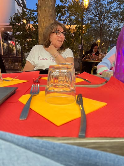 The scene is an outdoor restaurant setting in Montélimar, France, as suggested by the signage and context. The image is taken from the perspective of someone seated at a table. A woman with brown hair, wearing glasses, silver bracelets, and a white T-shirt with the word "Ciao" written on it, is looking at the camera. The table is covered with a red placemat and a yellow napkin. Silverware and a glass are placed on the table. In the background, another woman with long dark hair is seated at another table. There is a lamp post and trees visible in the background. The sky appears to be dusk or twilight.