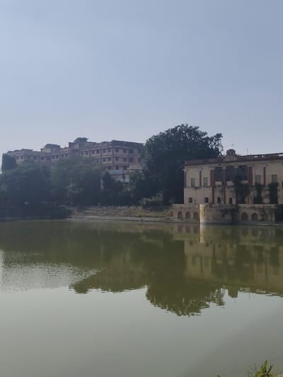 A tranquil pond reflects buildings on its edge and a distant structure on a hill. The building near the water has visible architectural elements like columns and arches. The reflection mirrors the architecture and nearby trees, creating a symmetrical effect. The water's surface is calm, contributing to the serene atmosphere of the scene. The sky is clear and light.