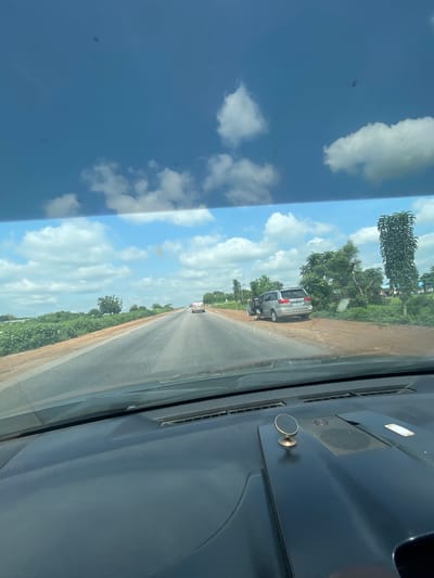 The image depicts a view from inside a vehicle traveling on a highway in Orei, Nigeria. The road ahead is paved and appears to be a two-lane highway with a dirt shoulder. On the right side of the road, there is a silver minivan parked with its driver's side door open. A person is partially visible near the open door, seemingly interacting with the vehicle. Further down the road, another car is visible. The surrounding environment consists of lush green vegetation and trees, characteristic of a rural or semi-rural area in Nigeria. The sky is bright blue with scattered white clouds. The foreground of the image shows the dashboard of the vehicle, including a magnetic phone mount. The scene captures a moment of travel on a Nigerian highway on a clear, cloudy day.