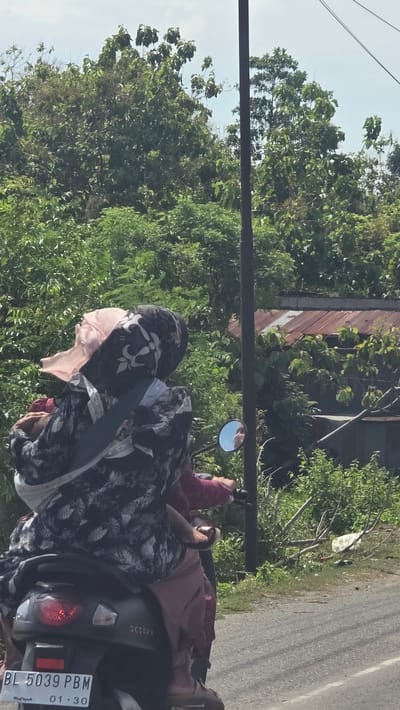 The image, taken in Teungoh Baro, Indonesia, shows two people on a black "Scoopy" scooter, located on an asphalt road. Lush green trees and vegetation line the background, with a building with a metal roof partially visible. A utility pole is situated on the left side of the image. The scooter's license plate reads "BL 5039 PBM" with an expiration date of "01.30". The individuals are dressed in clothing that covers their bodies, with the person in the front wearing a patterned scarf and the back passenger wearing a maroon outfit.