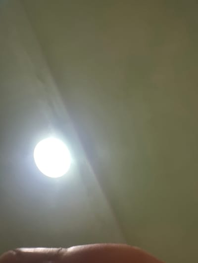 The image shows a bright, circular light source, possibly a ceiling light, on a light-colored, textured surface. The light source is positioned in the lower left quadrant of the image and emits a strong glow that diffuses outwards, creating subtle light streaks across the surface. The surface appears to be a wall or ceiling. In the very bottom of the frame, a blurred portion of a person's hand or finger is visible, casting a shadow. The overall impression is of an indoor environment, likely a room, with artificial lighting. There are no indications of specific landmarks or urban features that would suggest a location within the city of Corlăteni, Moldova.