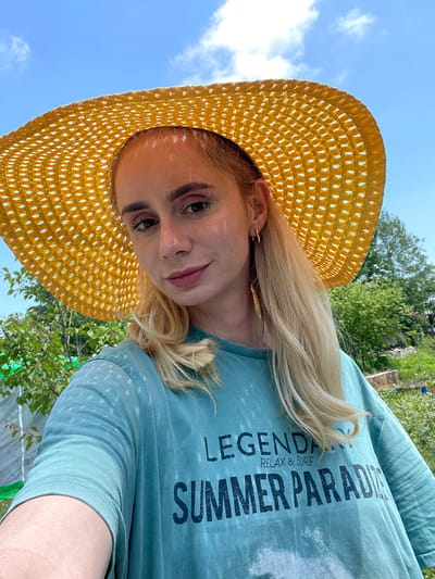 A young woman with blonde hair and fair skin is pictured in a close-up shot, wearing a large, yellow, woven sun hat and a light blue t-shirt. The t-shirt has text printed on it, with the words "LEGENDARY RELAX & SURF SUMMER PARADISE" visible. She has light makeup on and is wearing a small hoop earring in her visible ear. The background suggests an outdoor setting with lush green trees and foliage, under a clear blue sky with scattered clouds. The overall scene appears to be taken in a natural or garden environment. There is no indication of the location being the city of Knevichi, Russia.