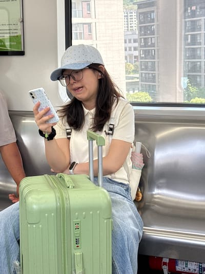 A young woman with long brown hair and glasses is seated in a train carriage with a light green suitcase beside her. She wears a white top, light blue jeans, and a blue polka-dot cap. She is holding a smartphone with a light blue polka-dot case in her left hand and looking at it. She has a black scrunchie on her wrist and a smartwatch on her other wrist. The suitcase has a combination lock set to "101" and "666." Through the train window, buildings are visible. A sign with text is partially visible on the left wall of the train. A fire extinguisher is mounted on the wall behind the woman. Another person's arm and shirt are also visible to the side of the woman, suggesting they are seated together on the train. The setting is suggestive of a train travelling through the city of Tiaodeng, China.