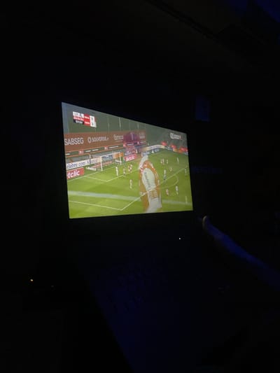 A low-lit room displays a soccer game on a screen. The broadcast shows a green field and players in red and white uniforms. On the lower left corner of the screen, match statistics are visible. An obscured leg is present in the upper right, suggesting someone is watching the game. Logos and advertisements line the field's perimeter on the display.