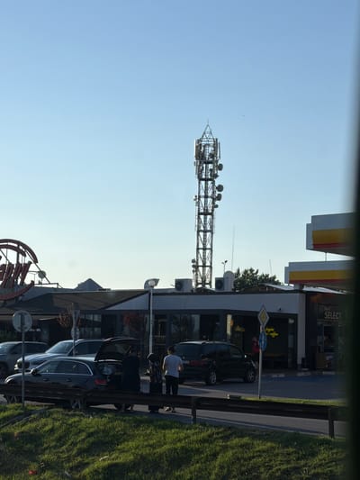 The image depicts a street scene in Radinovo, Bulgaria. A cell tower stands tall against a clear blue sky, equipped with multiple antennas. In the foreground, a dark-colored station wagon with its trunk open is parked, with several individuals gathered around it, seemingly loading or unloading items. Other vehicles, including a white SUV and a dark minivan, are also visible in the parking lot. A building with the "Shell Select" sign indicates a convenience store or gas station. The area is lined with a grassy verge and a black metal railing, suggesting it is along a road. The overall scene is captured in daylight, with sunlight casting shadows.