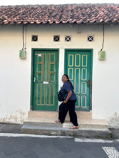 A woman is posing for a photo in front of two identical green doors with yellow trim, set against a white wall. The building has a tiled roof with a wave pattern. There are two square vents above each door. The woman is wearing a dark blue shirt, black pants, and brown sandals. She is carrying a black backpack and has a watch on her left wrist. She is smiling and looking towards the camera, with her body angled to the left. The doors are on a raised platform with a staircase leading up to them. The wall of the building shows signs of peeling paint. The ground in front of the building is paved with dark grey tiles. This image was taken in Banguntapan, Indonesia.