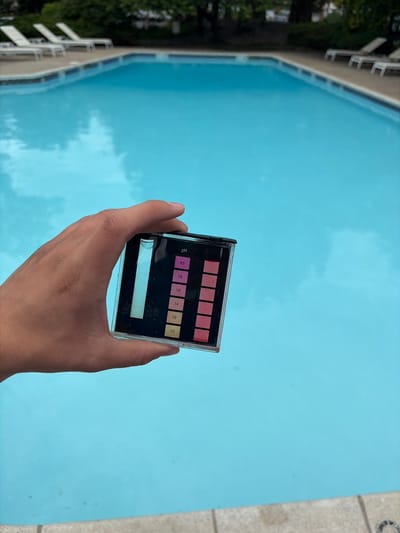 A person's hand holds a pool water testing kit above a swimming pool. The test kit has a color scale for pH levels, ranging from 7.0 to 8.0. The swimming pool is turquoise. Trees are visible in the upper left. The location is Edison, United States of America.