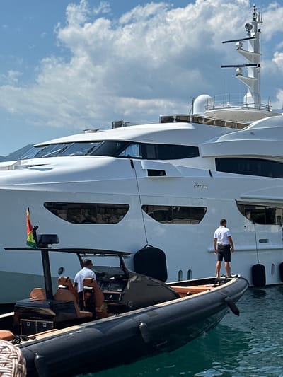 In Donja Lastva, Montenegro, a large white yacht is docked in the harbor. The yacht features multiple levels and numerous windows, with a nameplate visible on its hull. A man in a white polo shirt and black shorts stands on the yacht's deck, facing away from the camera. In the foreground, a dark-colored inflatable boat is tied to the yacht. Two individuals are visible inside the inflatable boat; one is standing and the other is seated. A flag, red, yellow, and green, is attached to a pole on the inflatable boat. The sky is partly cloudy, and the water appears calm.