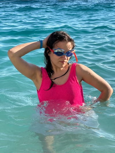 A young girl or woman is depicted floating on her back in clear, bright turquoise water, likely in the sea or a large pool, under direct sunlight. She wears a vibrant, textured fuchsia pink one-piece swimsuit and blue and orange swimming goggles. Her dark hair is wet, and her arms are bent with her hands resting near her head. A blue wristband is visible on her left wrist. The water's surface shows glistening ripples and splashes, particularly around her arms and torso, indicating motion or recent activity. She looks towards the viewer with a relaxed expression. The blue and orange goggles feature a fun shark tooth design on the sides, and partially visible text on the orange strap reads "DECATHLON TR". The bright sunlight illuminates the scene, enhancing the vivid colors of the water and the swimsuit. The context provided indicates the location is Kaş, Turkey.
