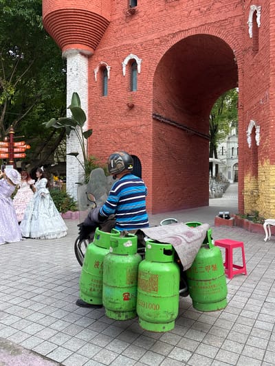 A man on a moped laden with green propane tanks is parked on a tiled street in Baqiao, China. Beside the moped is a trio of people in ornate Victorian gowns. In the background is a series of unusual brick buildings including an arched tunnelway, a cylindrical structure with horizontal ridges and small window features, and what appears to be a tourist information kiosk. The propane tanks are labelled with Chinese characters.