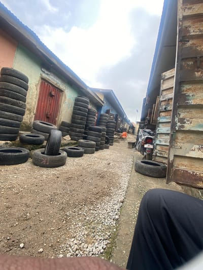 This is a forensic, matter-of-fact description of a scene in Pasere, Nigeria. The image depicts an outdoor scene, possibly a used tire shop or a roadside market. Stacks of used tires are prominently displayed on the left side of the image. A building with a weathered, painted facade, featuring a red door, is visible behind the tires. To the right, another building constructed with corrugated metal and weathered wood panels stands. A motorcycle is parked in front of this building, covered with a dark tarp. The ground is a mixture of dirt and gravel. The sky is overcast. A person's legs, clad in dark trousers, are visible in the lower right corner of the frame, suggesting the photographer's presence. The overall environment appears to be an urban or semi-urban area within Pasere, Nigeria.