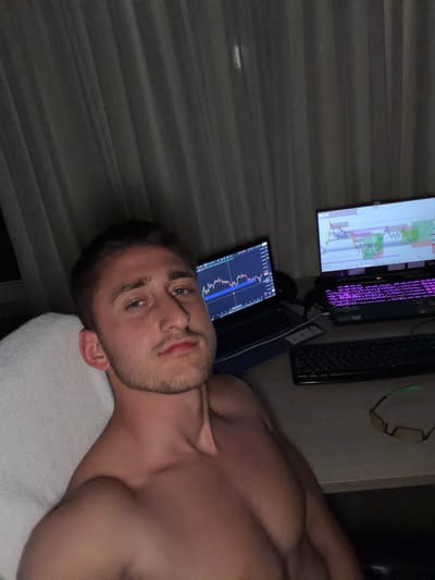 A fair-skinned man with short brown hair and a trimmed beard is lying down on a white surface, possibly a towel or pillow. He is shirtless, revealing his chest and shoulders. Behind him on a desk are two computer monitors, a laptop, and two keyboards. The screens display graphs, likely related to financial markets or trading. A pair of glasses rests on the desk as well. The location in Ede, Netherlands is not discernible from the image.