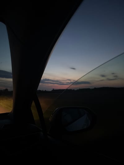The image captures a view from inside a vehicle, looking out the window during dusk. The sky displays a gradient of deep blue at the top transitioning to lighter blues and pinks near the horizon, with scattered clouds. A dark silhouette of trees and a distant landscape is visible across the middle ground. The foreground is dominated by the dark interior of the car, including a side mirror which reflects a portion of the car's interior, possibly a seat or headrest. To the left, a dark pillar, likely part of the car's frame, frames the view. A bright light source, possibly headlights or streetlights, is visible in the far left of the image, illuminating a portion of the road or surrounding area. The overall mood is serene and indicative of travel during twilight. No specific landmarks identifying Hajnówka, Poland, are discernible.