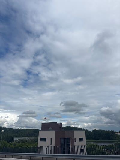 The image depicts a modern building situated in a natural landscape, under a dramatic, cloudy sky. The building features a contemporary design with contrasting materials, including light-colored plaster and dark wood paneling. It has large windows and a flat roof, on which a small red and black flag is flying. In front of the building, there is a metal fence and dense green trees. Beyond the trees, a body of water, likely a lake or river, is visible, with a forested hillside forming the backdrop. The sky is filled with a variety of clouds, ranging from light and wispy to dark and heavy, suggesting an overcast or approaching storm. The overall scene appears to be in a suburban or rural setting, characterized by the juxtaposition of man-made architecture and abundant nature. The location is identified as being within the Kapyĺski sel'ski Saveŭ, Belarus.