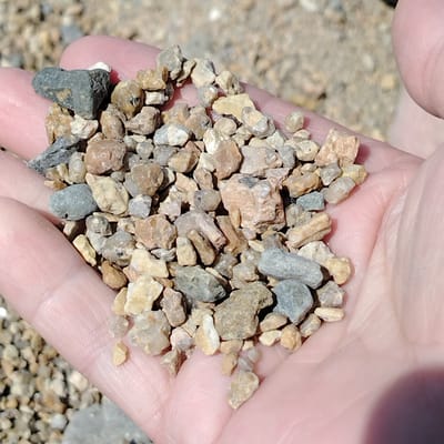 A close-up, over-the-shoulder shot shows a person's open palm filled with a small pile of gravel. The gravel consists of various sizes and colors of rocks, ranging from light beige and tan to darker gray and brown. Some pieces are angular, while others are more rounded. The hand holding the gravel is pale pink and appears to be part of a larger hand, with visible finger creases and texture. The background is out of focus, showing more gravel and a blurred, sandy-looking surface. The lighting suggests a bright, outdoor environment. The image was taken in the vicinity of Toraygyrov University in Toraygyrov, Kazakhstan.