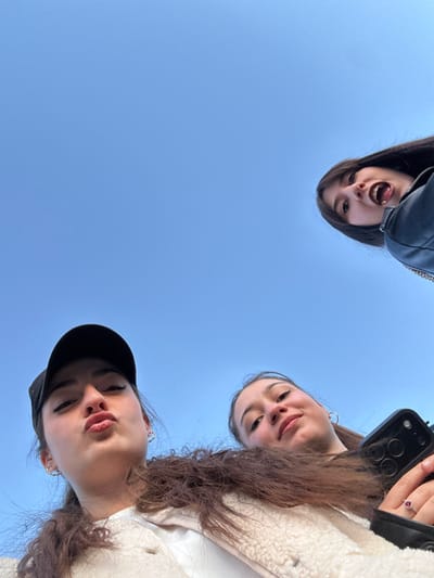 Three young women pose from a low angle against a clear blue sky. The woman on the left sticks her tongue out. The woman on the right holds a phone. The bottom woman wears a black baseball cap and has pursed lips. All three have light skin and long hair.