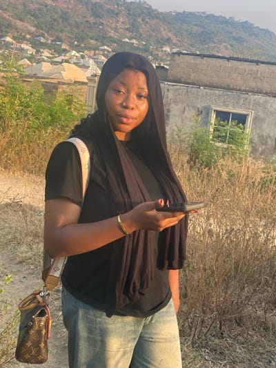 A young woman with a nose ring and a black head covering stands outdoors, holding a phone. She is wearing a black shirt, jeans, and a gold bracelet. A designer purse hangs from her shoulder. The background shows a hillside with buildings and dry vegetation. A building with a window is directly behind her. The image has been rotated.