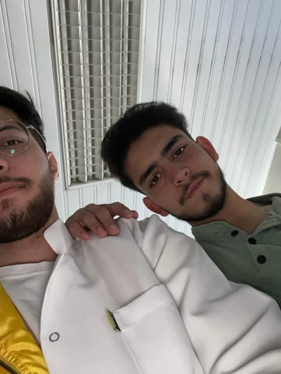 This image captures two young adult males in a close-up, low-angle shot, likely a selfie, taken indoors in Homs, Syria. The male on the left is partially visible, showing his left profile from the nose down, a dark beard, and round, light-framed eyeglasses. He wears a white t-shirt under a yellow garment, and a white collared jacket or shirt with a small yellow and black tag on a breast pocket, secured by a silver eyelet/rivet. The male on the right is centrally positioned, facing the camera with a direct gaze, displaying dark hair, a mustache, and a short beard. He wears a light green collared shirt with a placket featuring three dark buttons. His left hand rests on the left male's right shoulder. The overhead environment consists of a white or light-colored ceiling with parallel decorative strips and a rectangular fluorescent light fixture containing multiple tubes and a grid diffuser.