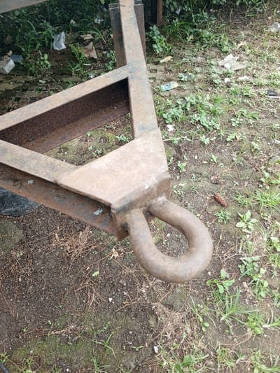 A close-up shot presents a rusty metal frame, likely part of a trailer hitch or similar apparatus. The frame is constructed of square tubing and features a large, rounded tow loop at its end. The metal shows signs of significant rust and wear. The surrounding ground is covered in dirt, weeds, and scattered debris like pine cones and trash. The image composition focuses primarily on the metal frame and its immediate environment, suggesting a neglected or discarded piece of equipment.