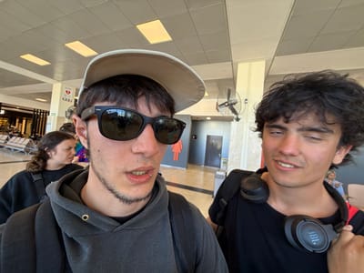 Two young men pose for a selfie-style shot in what appears to be an airport or transit hub. The man on the left has dark, wavy hair and wears headphones around his neck. The man on the right sports a dark hooded sweatshirt, sunglasses, and a beige cap. In the background, there are people, tiled floors, and what seems like a restroom sign. The image is noticeably rotated.