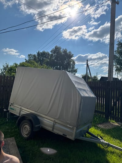 The image captures an outdoor scene in the village of Borodulino, Russia, on a sunny day with scattered clouds. In the foreground, a person's bare shoulder and back are visible, seated on a weathered wooden bench. The primary subject is a light-colored, tarp-covered trailer parked on a grassy area. The trailer is angled diagonally across the frame, with its wheels visible. A dark wooden fence runs along the mid-ground, partially obscured by the trailer and trees. In the background, the sky is a vibrant blue, with the sun shining intensely, casting lens flares and highlighting cumulus clouds. Electrical wires are strung across the sky, supported by a concrete utility pole on the right side of the image. The environment appears to be a rural or suburban setting, with trees and hints of distant buildings visible behind the fence.
