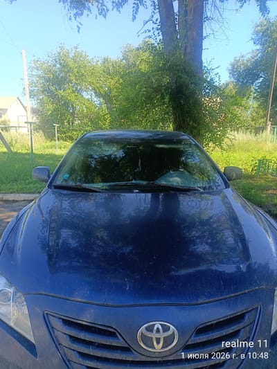 A dark blue Toyota sedan is parked outdoors in a residential area. The car's hood and windshield are visible, reflecting the surrounding environment of green trees, foliage, and a blue sky. A portion of a house with a light-colored roof and a wooden utility pole are also visible in the background, along with a decorative green fence. The scene appears to be in Aksay, Kazakhstan, as indicated by the text overlay on the image. The lighting suggests it is daytime, likely in the morning or afternoon, with shadows cast by the trees. The car's surface shows dust and some water spots.