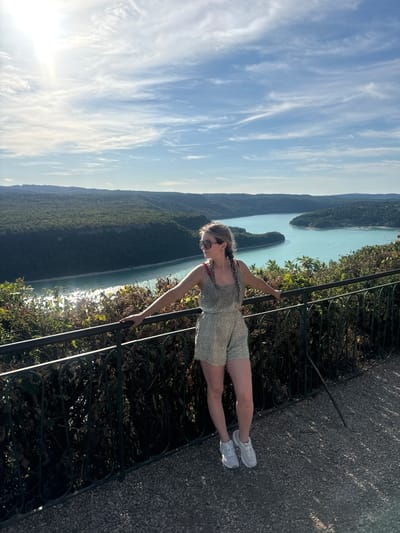 A young Caucasian woman with blonde hair in two braids, wearing a patterned romper and white sneakers, stands with her arms resting on a black metal railing. She is looking off to her left, with a scenic backdrop of a turquoise lake, lush green forested hills, and a blue sky with wispy clouds. The foreground shows a gravel path. The location is the Belvedere de La Forge in Moirans-en-Montagne, France, offering a panoramic view of the Lac de Vouglans.