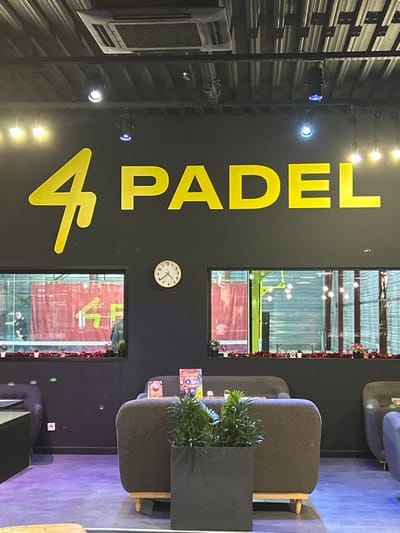 A brightly lit interior features the words "4 PADEL" in large yellow letters on a dark wall. Two windows showcase a padel court with players. A white clock is mounted on the wall. To the right, there are lounge chairs with small tables holding leaflets, along with a tall, dark planter with green plants. An air conditioning unit is visible on the left wall. The overall impression is a modern, stylish padel club reception area.