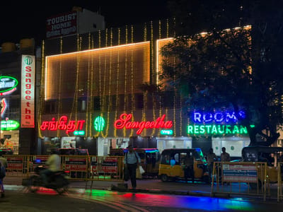 This is a nighttime, medium-angle shot of a street scene in Chennai, India, likely in a commercial area. The main focus is on a brightly lit restaurant called "Sangeetha Veg Restaurant," adorned with numerous yellow string lights cascading down its facade. Above "Sangeetha," a prominent neon sign spells out "ROOMS RESTAURANT" in blue and green. To the left, another restaurant's sign, "Bhavani Veg Restaurant," is partially visible.
The scene depicts a lively street with several auto-rickshaws parked and one in motion, blurred due to the camera's exposure. A few people are visible: one man in a light blue shirt walks with bags, another stands near an auto-rickshaw, and two more are inside one of the vehicles. Yellow traffic barriers are set up, with signs indicating "GREATER CHENNAI TRAFFIC POLICE" and "SILK SARES." The wet street reflects the colorful neon lights of the restaurants and vehicles, suggesting it may have recently rained. The overall atmosphere is vibrant and bustling, typical of a city street at night. The text "TOURIST HOME" is also visible on a sign above the restaurant, indicating lodging is available nearby.