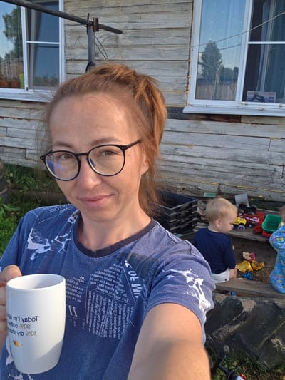 A medium shot shows a woman with red hair pulled back, wearing glasses and a blue t-shirt with white text, holding a white mug. She is outdoors, with a rustic wooden building behind her. The building has two windows, one with a curtain. To the right of the woman, a young child with blond hair, wearing a dark blue shirt and white shorts, is sitting in a sandbox playing with toys. Another child, wearing a blue patterned outfit, is partially visible in the sandbox. The environment appears to be rural, possibly a backyard, with grass and an old tire visible in the foreground. The overall lighting suggests it is daytime, possibly late afternoon. The location is identified as Буренка, Russia.