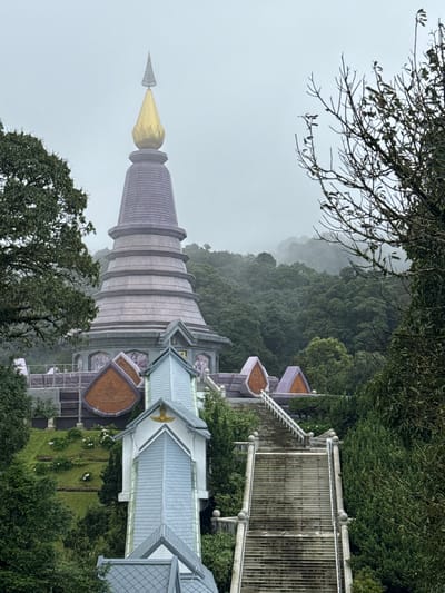 This image captures a serene, misty scene at a temple complex, likely Doi Inthanon National Park in Chang Khoeng, Thailand. The focal point is a tall, conical stupa with a striking golden spire, its pastel purple tiers fading into the overcast sky. To the right of the stupa, a white and grey building with a multi-layered roof and ornate eaves is visible. A long, elevated walkway with a metal railing and vertical slats runs alongside this building, suggesting an area for visitors to traverse or admire the scenery.
The surrounding landscape is lush and verdant, with dense green trees and foliage framing the structures. In the background, rolling hills disappear into the mist, creating a sense of depth and tranquility. The weather appears to be cool and damp, with a heavy fog obscuring the distance and lending a mystical atmosphere to the scene. The time of day is indeterminate due to the overcast conditions, but the soft light suggests it might be early morning or late afternoon.
No people are visible in the image, emphasizing the peaceful and contemplative nature of the location. The architecture, particularly the tiered stupa and the building's rooflines, points to a Buddhist temple setting. The overall mood is one of quiet beauty and spiritual calm, enhanced by the natural elements and the subdued lighting. There is no visible text in the image.