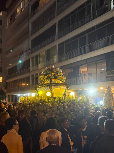 At night in Ancón, Peru, a religious procession proceeds along a street lined with buildings. People are gathered along the street. A lit float decorated with flowers and lights is carried by men in dark suits. The buildings are several stories high with visible windows.