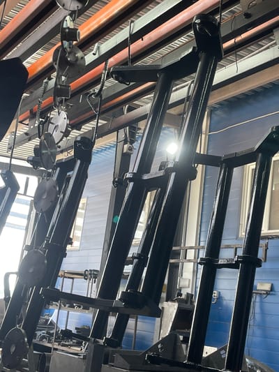In a workshop in Сокуры, Russia, multiple large black metal components, likely part of an industrial machine or structure, are suspended from an overhead track system. The components are cylindrical and angular, with various attachment points and visible welds. They appear to be either newly manufactured or undergoing maintenance, as they are coated in a shiny black finish, possibly paint or a protective sealant. The background shows a blue-paneled wall and industrial shelving, with fluorescent lighting casting a bright glow and creating reflections on the metal surfaces. The overall impression is one of an active manufacturing or repair facility.