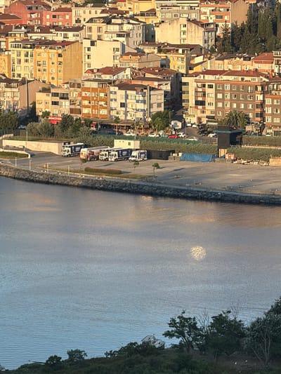 The image captures a waterfront scene in Eyüpsultan, Turkey, overlooking a body of water with a cityscape in the background. In the foreground, the calm water reflects the sunlight, creating a shimmering effect. Along the waterfront, a stone embankment separates the water from a paved area where several white trucks and trailers are parked. These vehicles appear to be transport trucks, some with logos visible. Beyond the trucks, a dense cluster of multi-story buildings with various colored facades and numerous windows forms the urban backdrop. The buildings exhibit a mix of architectural styles, with some featuring red tile roofs. Trees and vegetation are interspersed within the urban landscape and along the water's edge. The overall scene suggests an active port or industrial area within a densely populated city.