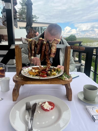 A man is eating a large meal presented on a wooden stand at a table in what appears to be an outdoor restaurant. The meal consists of multiple skewers of meat wrapped in bacon, served above a plate of assorted vegetables. To the right of the wooden stand are a plate with a sliced tomato topped with crumbled cheese, a fork and knife, a grey cup and saucer, and a white cigarette pack. The table is covered with a white tablecloth. The man is wearing a grey jacket. In the background, trees, buildings, and the sky are visible. The location is likely in the Smederevska Palanka Municipality, Serbia.