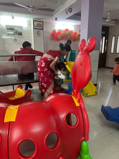 A woman in a maroon saree holds a baby on her lap near a stainless steel railing. A man in a red shirt stands nearby. A red and yellow playground structure dominates the foreground. A yellow and blue slide and a person standing are visible in the background. Wall decorations, a ceiling fan, and a balloon arrangement are also present. The image is rotated counterclockwise.