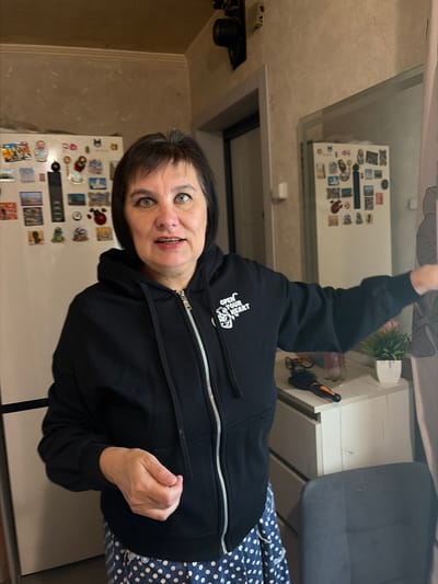 In a domestic kitchen setting in Tambov, Russia, a middle-aged woman with dark hair and green eyes stands in front of a refrigerator adorned with numerous stickers and magnets. She is wearing a black zip-up hoodie with the white text "OPEN YOUR HEART" on the left chest and a blue and white polka-dot skirt. The woman's right hand is held in front of her body, partially obscuring her view. To her right, a white dresser holds a small potted plant and a hairbrush. A chair is visible in the foreground. The lighting suggests it is daytime, with a light source from the left.