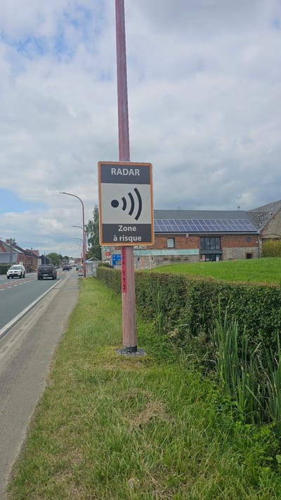 A radar speed camera sign is mounted on a red pole by the side of a road in Boussu-lez-Walcourt, Belgium. The sign is rectangular with an orange border and a white background. It displays the word "RADAR" in black letters at the top, followed by a black radar symbol and the text "Zone à risque" (Risk zone) in black. A white arrow pointing left with "Deneu" and an arrow pointing up with "Boussu" are visible on a smaller blue sign below the radar sign. Red markings, E206 and 113, are visible vertically on the pole. The road is paved and has a white line marking. Several vehicles, including a white car and a dark SUV, are driving on the road. A building with a brick facade and solar panels on the roof is situated behind a green hedge. The environment is characterized by green grass and vegetation on the right side of the road. The sky is cloudy with patches of blue.