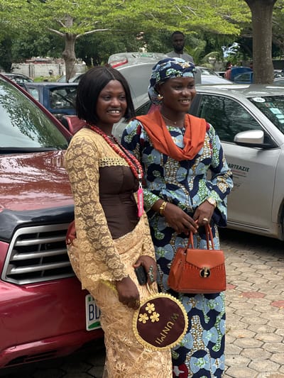 In the city of Itoga, Nigeria, two women are standing next to a red vehicle. The woman on the left is wearing a gold lace overlay over a brown dress, a red beaded necklace, and a brown handbag. She is holding a decorative circular fan with gold floral embellishments and the word "WUNMI" written in gold lettering. The woman on the right is wearing a blue, white, and green patterned dress with an orange headscarf and shawl. She is holding an orange textured handbag. A man is visible in the background, standing behind some cars. The environment appears to be an outdoor parking area, with trees and paved ground.