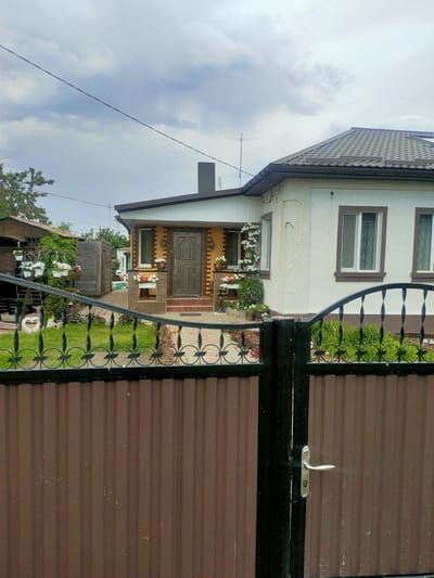 This image depicts the entrance to a private residence in Nosivka, Ukraine, on a cloudy day. A tall, corrugated brown metal gate with a black decorative wrought-iron fence tops it, and a silver door handle is visible on the right side of the gate. Beyond the gate, a pathway leads to a house with white stucco walls and brown trim around the windows and doors. The main entrance is a dark brown door with decorative carvings, framed by two amber-colored wall sconces. Several flower boxes with white and red flowers adorn the front of the house. To the left of the main house, a smaller, yellowish structure with a dark brown door is visible, also decorated with flower boxes. A wooden fence and more greenery can be seen further back. The scene suggests a well-maintained residential property.