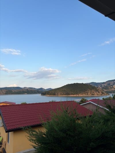 The image shows a scenic view of a lake with rolling hills and mountains in the background. In the foreground, there are red-tiled roofs of houses and green evergreen trees. The sky is clear blue with a few scattered white clouds. The scene appears to be a tranquil, natural landscape with some residential development.