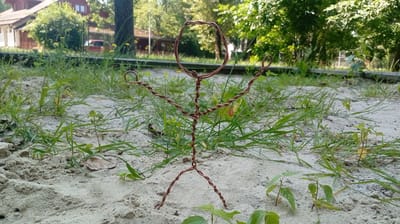 A wire figurine of a stick figure stands in a sandy area surrounded by grass. The figurine is made of copper wire and has a circular head, a twisted body, and bent arms and legs. The setting appears to be outdoors, with trees and a building in the background. The time of day seems to be daytime, likely sunny, given the lighting. The figurine is positioned in the center of the frame, with the sand and grass in the foreground and the background blurred. There is no visible text in the image. The overall mood is whimsical and playful, suggesting a handmade creation placed in a natural environment. The location context of Karviná, Czechia, indicates the geographical setting of this scene.